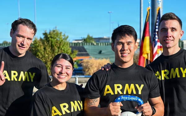 Four U.S. Army personnel pose together outdoors in matching black Army PT (Physical Training) shirts and shorts. One individual holds a skull-shaped trophy or award labeled 'PERSHING,' and another gives a thumbs-up gesture. The group appears to be celebrating a competition or event victory, with U.S. and German flags visible in the background.
