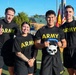 Four U.S. Army personnel pose together outdoors in matching black Army PT (Physical Training) shirts and shorts. One individual holds a skull-shaped trophy or award labeled 'PERSHING,' and another gives a thumbs-up gesture. The group appears to be celebrating a competition or event victory, with U.S. and German flags visible in the background.