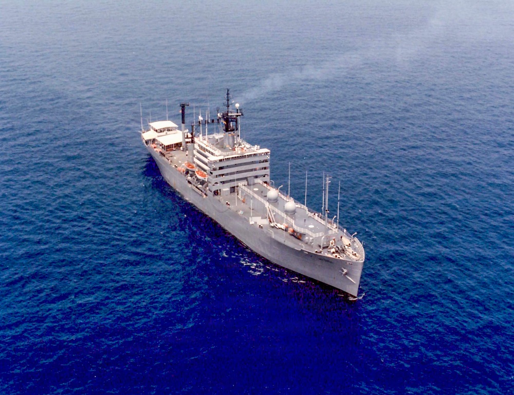 An aerial photograph of a gray military or government vessel underway in open blue ocean waters. The ship appears to be a mid-sized auxiliary or research/command vessel with multiple antenna masts, radar domes, and a multi-deck superstructure. Light smoke is visible from one of the stacks, indicating the engines are running.