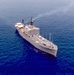 An aerial photograph of a gray military or government vessel underway in open blue ocean waters. The ship appears to be a mid-sized auxiliary or research/command vessel with multiple antenna masts, radar domes, and a multi-deck superstructure. Light smoke is visible from one of the stacks, indicating the engines are running.