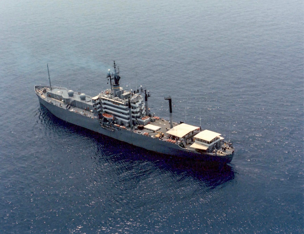 An aerial photograph of a gray military or naval auxiliary vessel underway on calm, open blue water. The ship features a prominent superstructure amidships with multiple decks, radar domes, masts, and antenna arrays, suggesting intelligence-gathering or command-and-control functions. The aft deck area includes what appears to be a large white canvas or tarpaulin covering, along with various deck equipment.