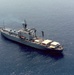 An aerial photograph of a gray military or naval auxiliary vessel underway on calm, open blue water. The ship features a prominent superstructure amidships with multiple decks, radar domes, masts, and antenna arrays, suggesting intelligence-gathering or command-and-control functions. The aft deck area includes what appears to be a large white canvas or tarpaulin covering, along with various deck equipment.