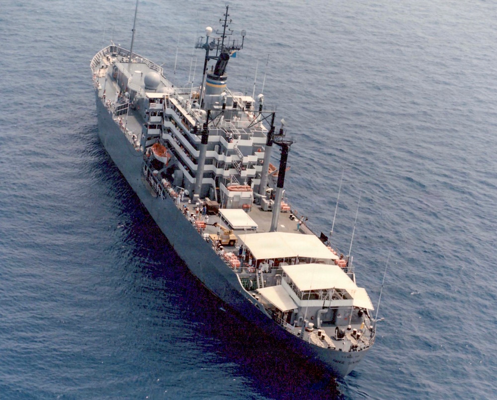 An aerial photograph of a large gray naval auxiliary or support vessel underway at sea. The ship features a prominent superstructure with multiple deck levels, various antennas and masts, and what appear to be replenishment or cargo handling structures. The image appears to be from the Cold War era based on photo quality and ship design.