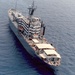 An aerial photograph of a large gray naval auxiliary or support vessel underway at sea. The ship features a prominent superstructure with multiple deck levels, various antennas and masts, and what appear to be replenishment or cargo handling structures. The image appears to be from the Cold War era based on photo quality and ship design.