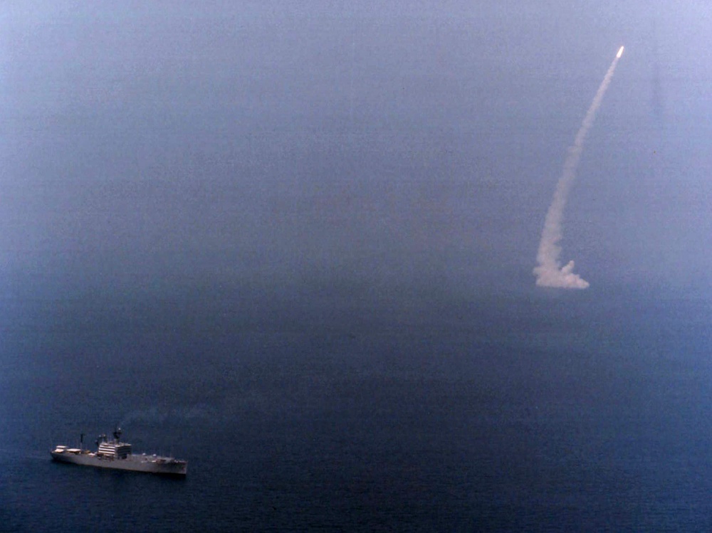 An aerial photograph shows a naval warship at sea with a missile or rocket visibly launched and ascending steeply into the sky, leaving a prominent smoke trail. The ship appears to be a mid-sized naval vessel positioned in the lower-left portion of the frame. The image captures what appears to be a live missile test or combat firing exercise at sea.
