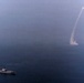 An aerial photograph shows a naval warship at sea with a missile or rocket visibly launched and ascending steeply into the sky, leaving a prominent smoke trail. The ship appears to be a mid-sized naval vessel positioned in the lower-left portion of the frame. The image captures what appears to be a live missile test or combat firing exercise at sea.