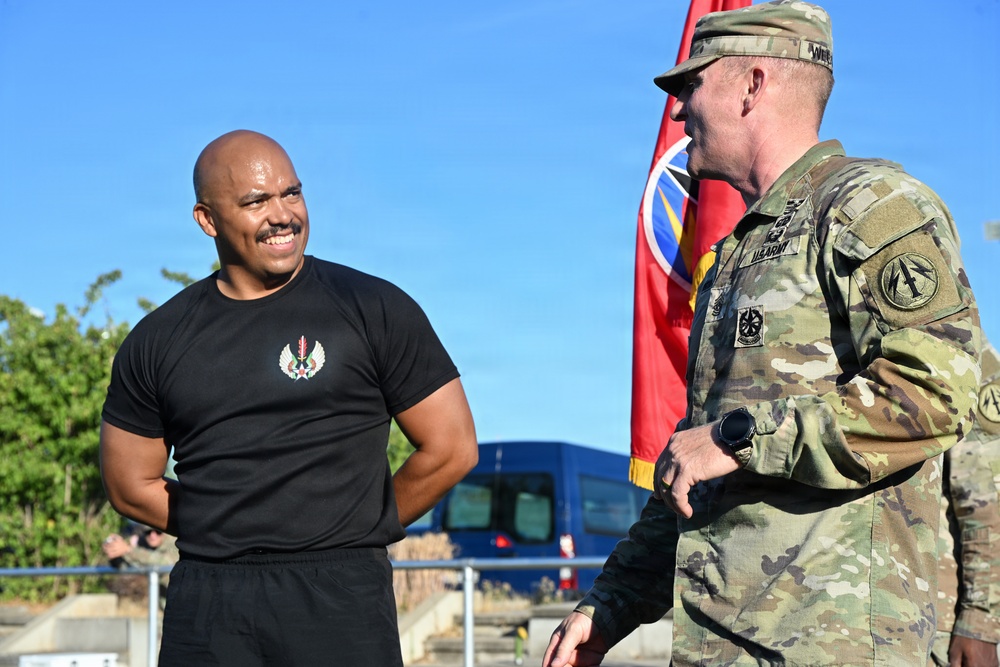 Two individuals are engaged in conversation outdoors, one wearing a black athletic t-shirt with an emblem and the other in U.S. Army MultiCam (OCP) camouflage uniform. A red military unit guidon or flag is visible in the background. The setting appears to be a military installation or outdoor event area.