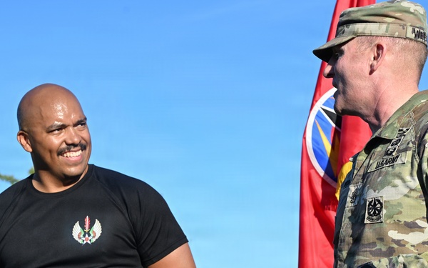 Two individuals are engaged in conversation outdoors, one wearing a black athletic t-shirt with an emblem and the other in U.S. Army MultiCam (OCP) camouflage uniform. A red military unit guidon or flag is visible in the background. The setting appears to be a military installation or outdoor event area.