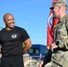 Two individuals are engaged in conversation outdoors, one wearing a black athletic t-shirt with an emblem and the other in U.S. Army MultiCam (OCP) camouflage uniform. A red military unit guidon or flag is visible in the background. The setting appears to be a military installation or outdoor event area.