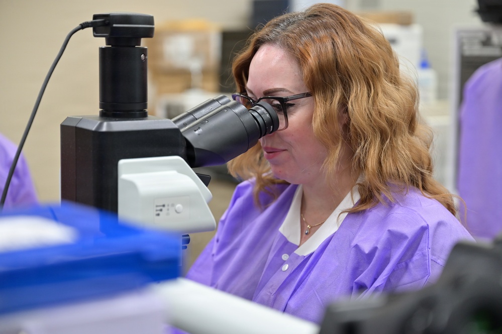 A woman wearing a purple lab coat and black-framed glasses is looking through the eyepiece of a microscope in a laboratory setting. The microscope is equipped with a camera attachment connected via a black cable. The background shows a typical lab environment with various supplies and equipment.
