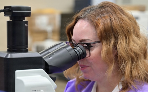 A woman wearing a purple lab coat and black-framed glasses is looking through the eyepiece of a microscope in a laboratory setting. The microscope is equipped with a camera attachment connected via a black cable. The background shows a typical lab environment with various supplies and equipment.