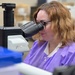 A woman wearing a purple lab coat and black-framed glasses is looking through the eyepiece of a microscope in a laboratory setting. The microscope is equipped with a camera attachment connected via a black cable. The background shows a typical lab environment with various supplies and equipment.