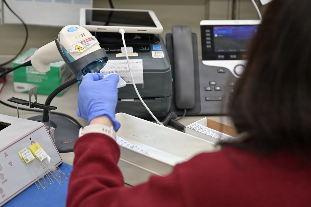A person wearing blue latex gloves and a red sweater is scanning a barcode label using a handheld barcode scanner at a workstation. The setting appears to be a laboratory, pharmacy, or medical/clinical environment based on the equipment and materials visible. The worker is processing labeled items, likely for inventory, specimen tracking, or medication management.