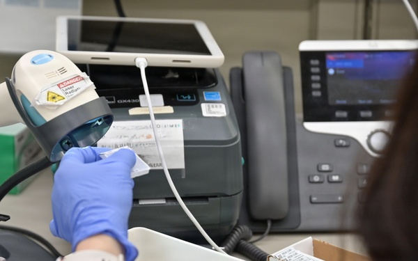 A person wearing blue latex gloves and a red sweater is scanning a barcode label using a handheld barcode scanner at a workstation. The setting appears to be a laboratory, pharmacy, or medical/clinical environment based on the equipment and materials visible. The worker is processing labeled items, likely for inventory, specimen tracking, or medication management.
