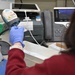 A person wearing blue latex gloves and a red sweater is scanning a barcode label using a handheld barcode scanner at a workstation. The setting appears to be a laboratory, pharmacy, or medical/clinical environment based on the equipment and materials visible. The worker is processing labeled items, likely for inventory, specimen tracking, or medication management.