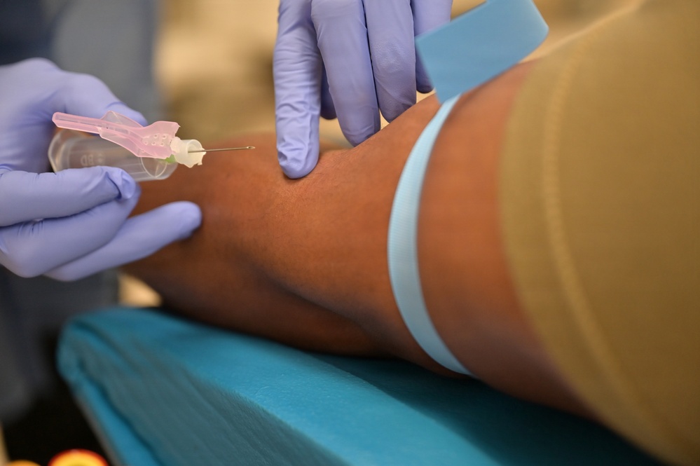 A medical professional wearing blue nitrile gloves is preparing to insert an intravenous (IV) catheter or perform a venipuncture on a patient's arm. A light blue tourniquet band is visible on the upper forearm to distend the vein. The needle device appears to be a standard IV cannula with a pink hub.