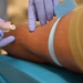 A medical professional wearing blue nitrile gloves is preparing to insert an intravenous (IV) catheter or perform a venipuncture on a patient's arm. A light blue tourniquet band is visible on the upper forearm to distend the vein. The needle device appears to be a standard IV cannula with a pink hub.