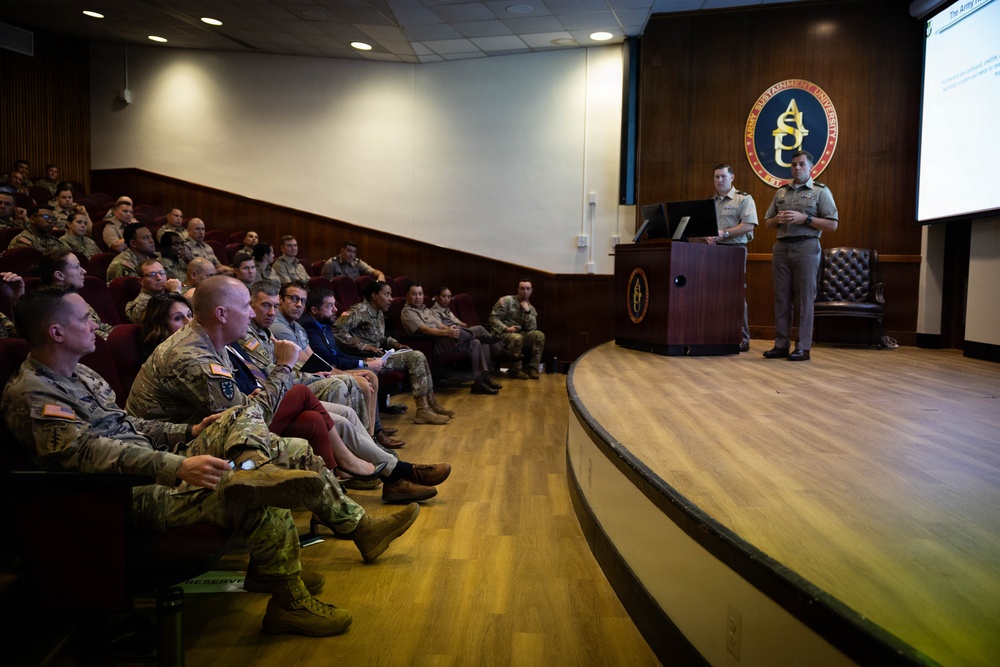 A military briefing or lecture is taking place in an auditorium at Army Sustainment University (ASU), as indicated by the seal visible on the back wall. Two uniformed military personnel stand at a podium on a raised stage, presenting to an audience of approximately 30-40 military and civilian attendees seated in red auditorium chairs. A projection screen is visible to the right displaying presentation content.
