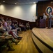 A military briefing or lecture is taking place in an auditorium at Army Sustainment University (ASU), as indicated by the seal visible on the back wall. Two uniformed military personnel stand at a podium on a raised stage, presenting to an audience of approximately 30-40 military and civilian attendees seated in red auditorium chairs. A projection screen is visible to the right displaying presentation content.