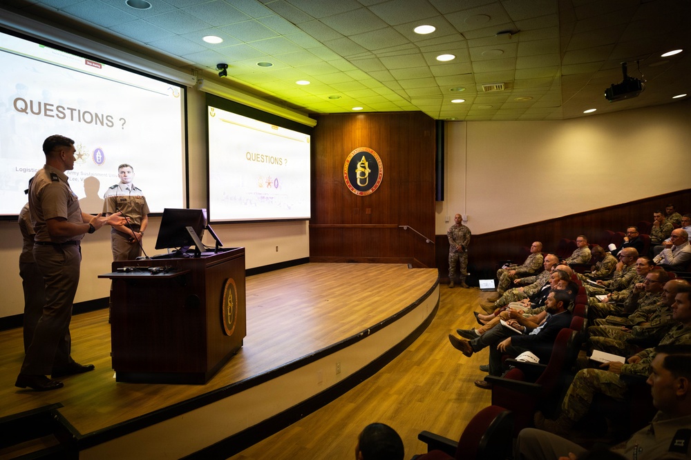A military briefing or presentation is taking place in an auditorium-style lecture hall, with two uniformed personnel presenting at a podium on a raised stage. The presentation slide displays 'QUESTIONS?' indicating the briefing is concluding. An audience of approximately 20-30 military and civilian personnel are seated in tiered seating.