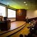 A military briefing or presentation is taking place in an auditorium-style lecture hall, with two uniformed personnel presenting at a podium on a raised stage. The presentation slide displays 'QUESTIONS?' indicating the briefing is concluding. An audience of approximately 20-30 military and civilian personnel are seated in tiered seating.