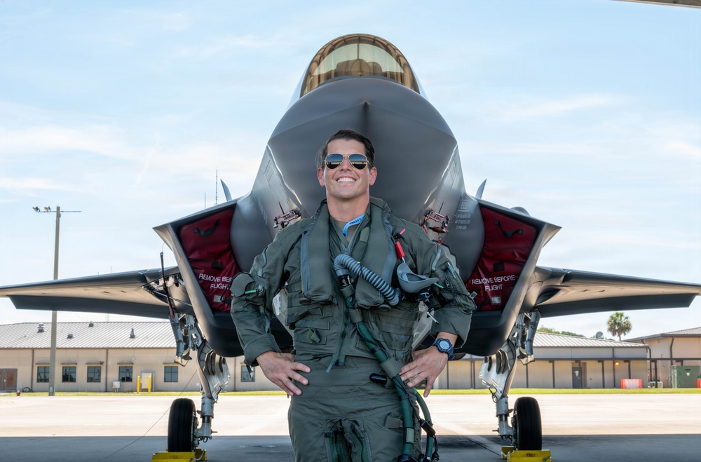 A military pilot in full flight gear stands confidently in front of an F-35 Lightning II stealth fighter jet on a tarmac. The pilot is smiling and wearing aviator sunglasses, an olive flight suit, and a survival vest with oxygen mask hose. The setting appears to be a military air base with hangars visible in the background.