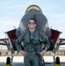 A military pilot in full flight gear stands confidently in front of an F-35 Lightning II stealth fighter jet on a tarmac. The pilot is smiling and wearing aviator sunglasses, an olive flight suit, and a survival vest with oxygen mask hose. The setting appears to be a military air base with hangars visible in the background.