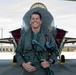 A military pilot in a green flight suit stands smiling in front of a modern fighter jet, identified by visible 'REMOVE BEFORE FLIGHT' tags on the aircraft. The setting appears to be a military air base with hangars and infrastructure visible in the background. The pilot is wearing full flight gear including a G-suit, harness, and oxygen mask assembly.