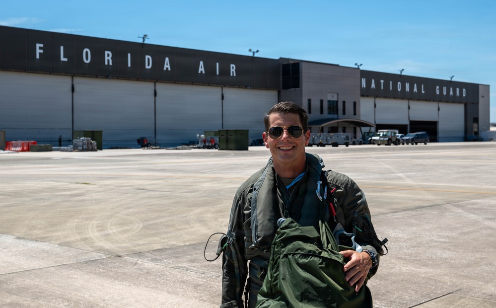A military aviator in a flight suit and sunglasses stands on an aircraft ramp in front of a large hangar bearing the text 'FLORIDA AIR NATIONAL GUARD'. The individual is smiling and carrying green flight gear, suggesting they have recently completed or are preparing for a flight. The setting is a military airfield operated by the Florida Air National Guard.