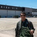 A military aviator in a flight suit and sunglasses stands on an aircraft ramp in front of a large hangar bearing the text 'FLORIDA AIR NATIONAL GUARD'. The individual is smiling and carrying green flight gear, suggesting they have recently completed or are preparing for a flight. The setting is a military airfield operated by the Florida Air National Guard.