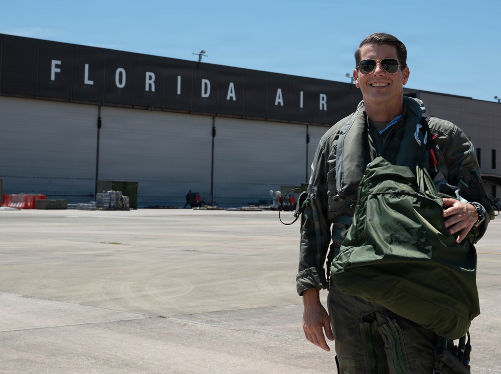 A military pilot in a flight suit and aviator sunglasses stands on an airport tarmac, smiling and holding a green parachute pack or flight bag. Behind him is a large hangar with 'FLORIDA AIR' written in bold white letters on a dark fascia. The setting appears to be a military or joint-use airfield in Florida under clear, sunny skies.