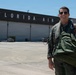 A military pilot in a flight suit and aviator sunglasses stands on an airport tarmac, smiling and holding a green parachute pack or flight bag. Behind him is a large hangar with 'FLORIDA AIR' written in bold white letters on a dark fascia. The setting appears to be a military or joint-use airfield in Florida under clear, sunny skies.