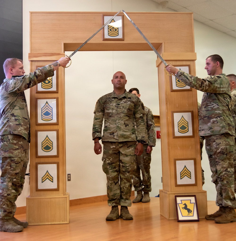 A U.S. Army NCO promotion or induction ceremony is taking place indoors, featuring a ceremonial arch of sabers. Two soldiers hold crossed sabers overhead while a central soldier in OCP (Operational Camouflage Pattern) uniform stands beneath the arch. The wooden arch displays NCO rank insignia plaques from Corporal through Sergeant Major, indicating this is likely an NCO induction ceremony.