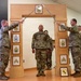 A U.S. Army NCO promotion or induction ceremony is taking place indoors, featuring a ceremonial arch of sabers. Two soldiers hold crossed sabers overhead while a central soldier in OCP (Operational Camouflage Pattern) uniform stands beneath the arch. The wooden arch displays NCO rank insignia plaques from Corporal through Sergeant Major, indicating this is likely an NCO induction ceremony.