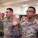 A group of U.S. Army soldiers are raising their right hands in what appears to be a re-enlistment or oath ceremony held indoors. The foreground shows two soldiers clearly identifiable by name tapes and rank insignia, with a larger formation of soldiers visible in the background. All personnel are wearing Army Combat Uniforms (ACU) in Operational Camouflage Pattern (OCP).