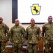 Four U.S. Army soldiers in OCP (Operational Camouflage Pattern) uniforms stand at attention during a formal military ceremony. A projection screen behind them displays a unit insignia featuring a rearing horse on a yellow keystone shape, consistent with Pennsylvania National Guard imagery. Additional personnel are visible in the background near a podium and stage area.