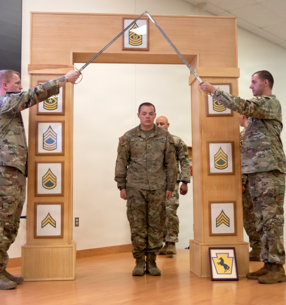 A U.S. Army NCO induction or promotion ceremony is taking place indoors, where a soldier named Allen is walking through a ceremonial arch while two soldiers on either side cross sabers overhead. The wooden arch displays NCO rank insignia plaques from Specialist through Sergeant Major of the Army. A Pennsylvania National Guard unit insignia framed print is visible at the base of the arch.