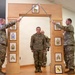 A U.S. Army NCO induction or promotion ceremony is taking place indoors, where a soldier named Allen is walking through a ceremonial arch while two soldiers on either side cross sabers overhead. The wooden arch displays NCO rank insignia plaques from Specialist through Sergeant Major of the Army. A Pennsylvania National Guard unit insignia framed print is visible at the base of the arch.