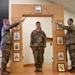 Three U.S. Army soldiers are participating in a formal NCO induction ceremony. Two soldiers holding sabers form an arch over a third soldier standing at attention beneath a decorative wooden arch displaying NCO rank insignia plaques. A framed unit crest (appears to be 28th Infantry Division) is visible on the floor at the base of the arch.