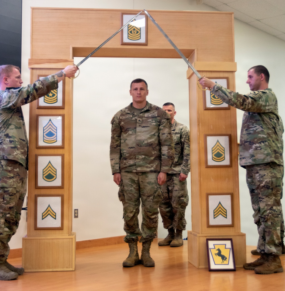 A U.S. Army NCO promotion or induction ceremony is taking place indoors. A soldier identified by nametape as 'ONYSHCHUK' stands at attention beneath a ceremonial arch while two soldiers cross sabers overhead. The wooden arch displays NCO rank insignia plaques from Specialist through Sergeant Major of the Army, and a unit crest is visible at the base.