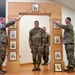 A U.S. Army NCO promotion or induction ceremony is taking place indoors. A soldier identified by nametape as 'ONYSHCHUK' stands at attention beneath a ceremonial arch while two soldiers cross sabers overhead. The wooden arch displays NCO rank insignia plaques from Specialist through Sergeant Major of the Army, and a unit crest is visible at the base.