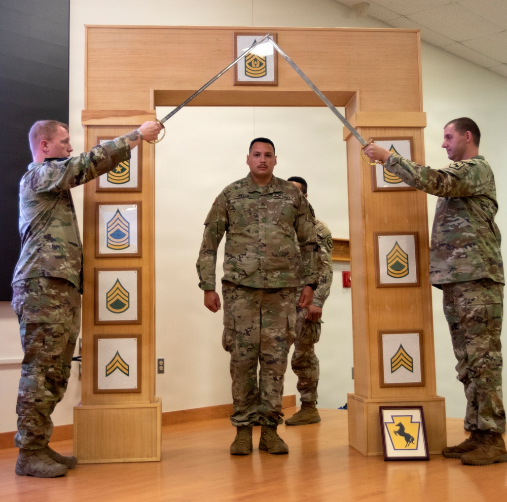 A U.S. Army NCO induction or promotion ceremony is taking place indoors. Two soldiers in OCP (Operational Camouflage Pattern) uniforms hold crossed sabers forming an arch, while a third soldier stands beneath them passing through. The ceremonial arch structure displays NCO rank insignia plaques from Corporal through Sergeant Major.