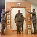 A U.S. Army NCO induction or promotion ceremony is taking place indoors. Two soldiers in OCP (Operational Camouflage Pattern) uniforms hold crossed sabers forming an arch, while a third soldier stands beneath them passing through. The ceremonial arch structure displays NCO rank insignia plaques from Corporal through Sergeant Major.