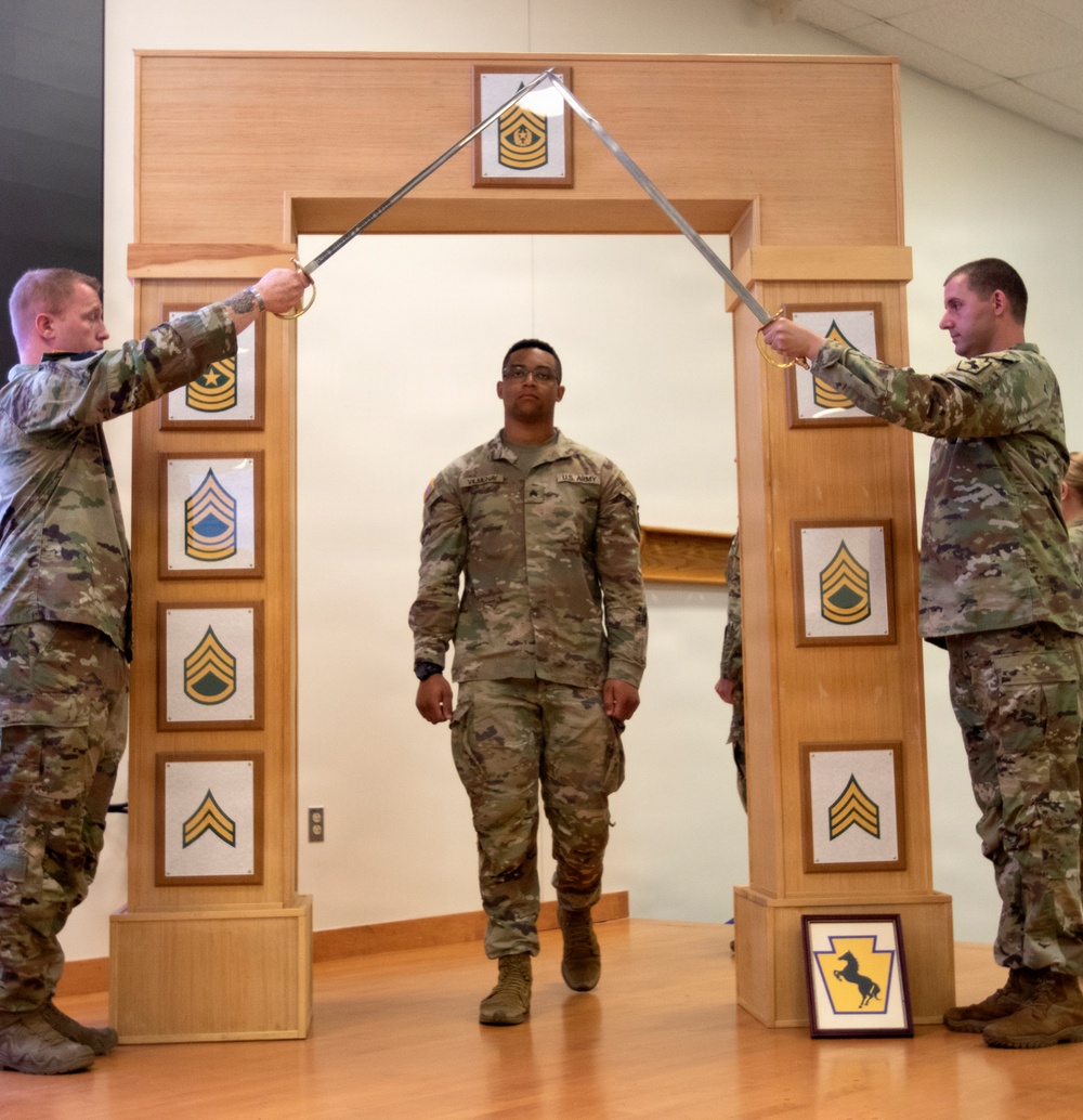 A U.S. Army NCO induction or promotion ceremony is taking place indoors, with a soldier walking through a ceremonial arch while two soldiers on either side cross sabers overhead. The wooden arch displays multiple NCO rank insignia plaques from Specialist through Sergeant Major of the Army. A framed unit crest (appearing to be a horse/mustang insignia) is visible on the floor at the base of the arch.