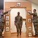 A U.S. Army NCO induction or promotion ceremony is taking place indoors, with a soldier walking through a ceremonial arch while two soldiers on either side cross sabers overhead. The wooden arch displays multiple NCO rank insignia plaques from Specialist through Sergeant Major of the Army. A framed unit crest (appearing to be a horse/mustang insignia) is visible on the floor at the base of the arch.