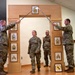 A U.S. Army NCO induction or promotion ceremony is taking place indoors, featuring a soldier walking through a ceremonial arch while two soldiers cross sabers overhead. The wooden arch displays NCO rank insignia plaques from Specialist through Sergeant Major of the Army. A framed unit crest (appearing to be a cavalry or Pennsylvania unit) is visible on the floor at the base of the arch.