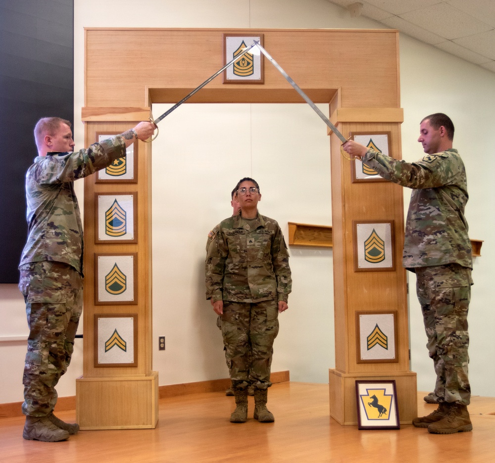 Three U.S. Army soldiers are participating in a formal NCO induction ceremony. Two soldiers on either side hold crossed sabers over a third soldier standing at attention beneath a decorative wooden arch displaying NCO rank insignia plaques. A framed unit patch is visible on the floor to the right.