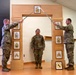 Three U.S. Army soldiers are participating in a formal NCO induction ceremony. Two soldiers on either side hold crossed sabers over a third soldier standing at attention beneath a decorative wooden arch displaying NCO rank insignia plaques. A framed unit patch is visible on the floor to the right.