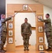 Three U.S. Army soldiers are participating in a formal NCO induction or promotion ceremony. Two soldiers on either side hold crossed swords over a ceremonial arch while a third soldier stands at attention beneath the arch. The wooden arch displays various NCO rank insignia plaques, and a unit crest is visible on the floor.
