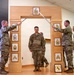 A U.S. Army promotion or graduation ceremony is taking place indoors, featuring a ceremonial arch with NCO rank insignia plaques displayed on its columns. Two soldiers are crossing sabers/swords over the arch while a third soldier stands beneath it at attention. A framed unit crest (appearing to be a horse/Pegasus insignia) is displayed on the floor in front of the arch.